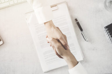 Zwei Personen geben sich die Hand, darunter ein Dokument auf einem Schreibtisch.