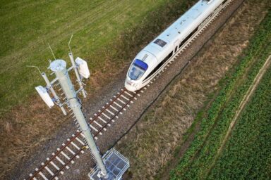 Es ist eine Bahn auf einem Gleis zu sehen, welche zwischen Wiese durchfährt. Im Vordergrund des Bildes ist ein Funkmast zu sehen.