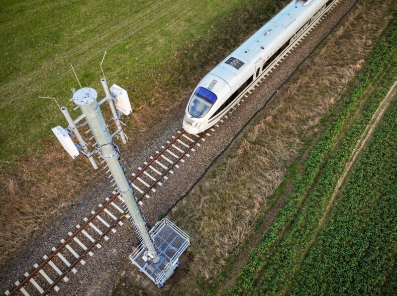 Es ist eine Bahn auf einem Gleis zu sehen, welche zwischen Wiese durchfährt. Im Vordergrund des Bildes ist ein Funkmast zu sehen.