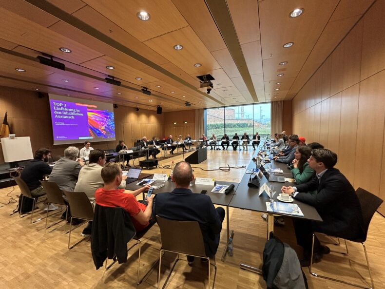 Die Teilnehmenden am Evaluierungstreffen sitzen in einem Besprechungsraum an einem großen Tisch. Auf einer Leinwand sieht man eine Power Point Präsentation.